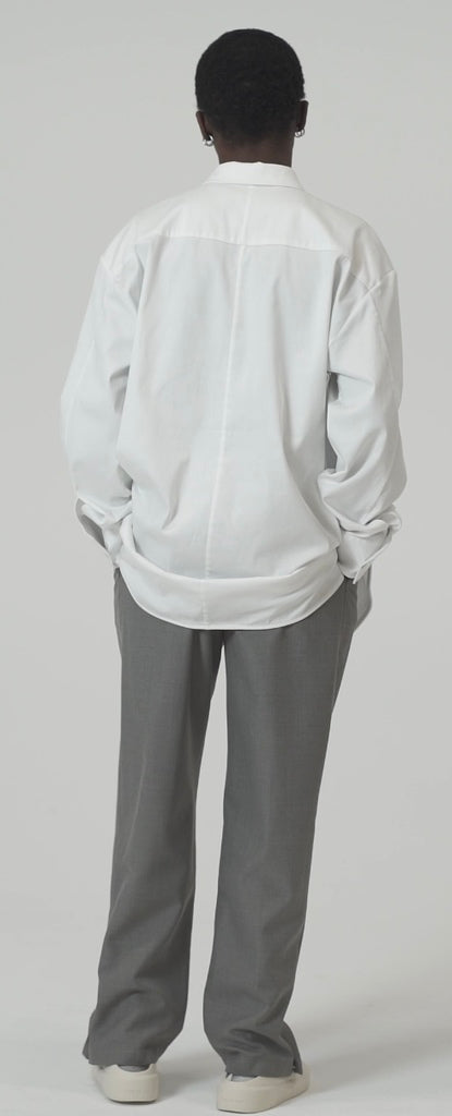 Model wearing an oversized cotton blend shirt in white, showcasing the back design and relaxed fit with gray pants.