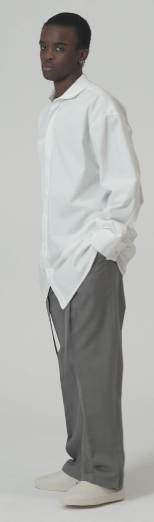 Model wearing an oversized cotton blend poplin shirt in white with an oversized pocket, paired with gray trousers.