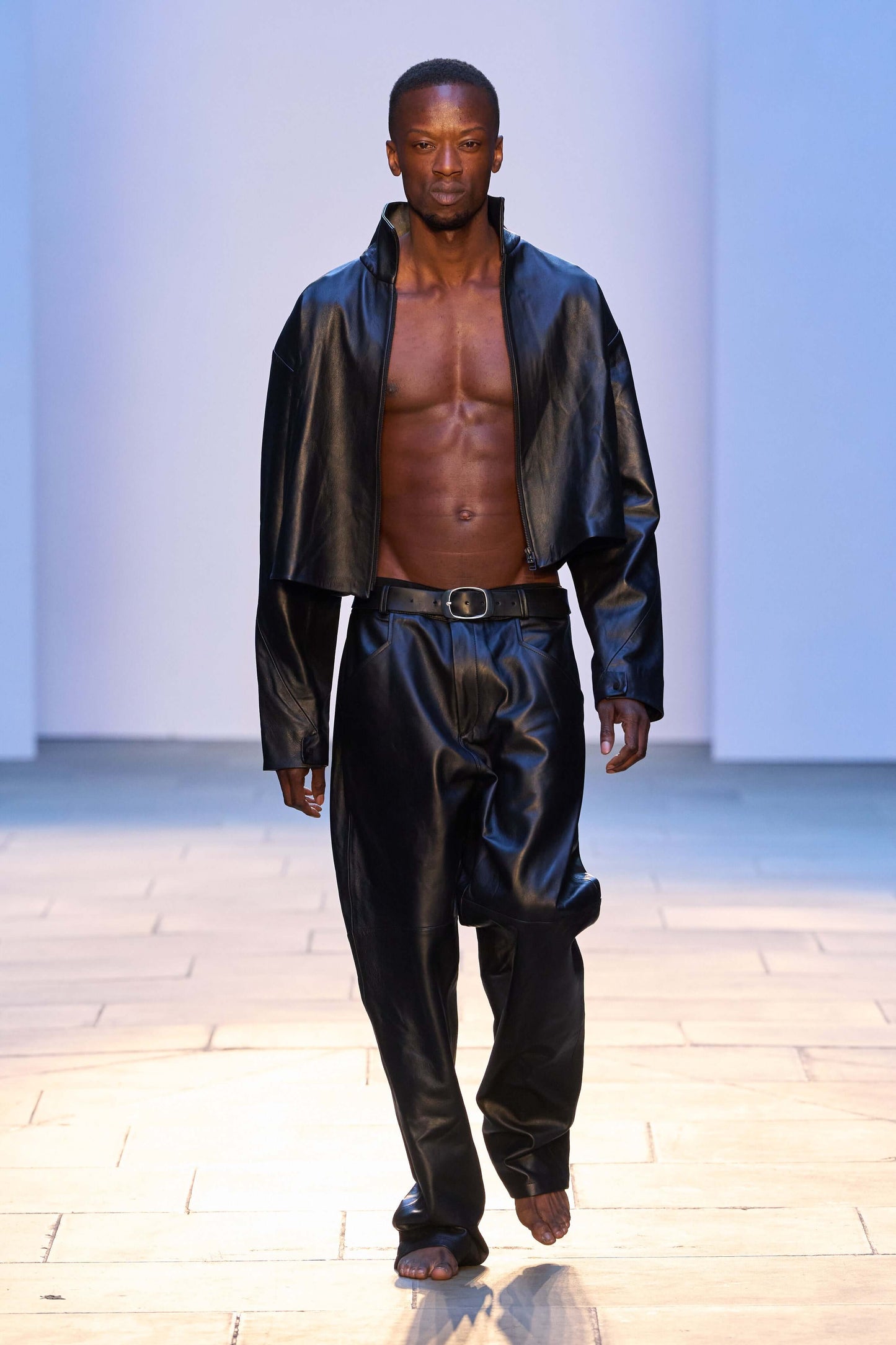Male model showcasing black leather trousers with a cropped jacket on a runway.