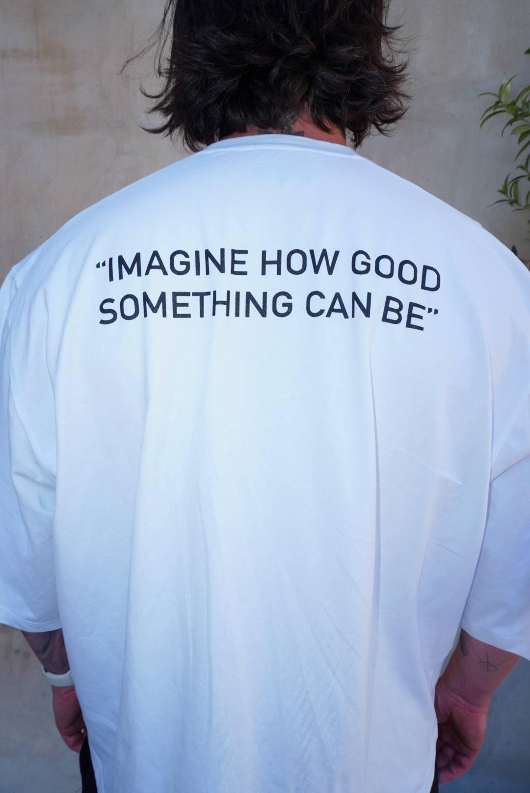 Back view of white boxy tee with black screen print, featuring the quote 'IMAGINE HOW GOOD SOMETHING CAN BE'.