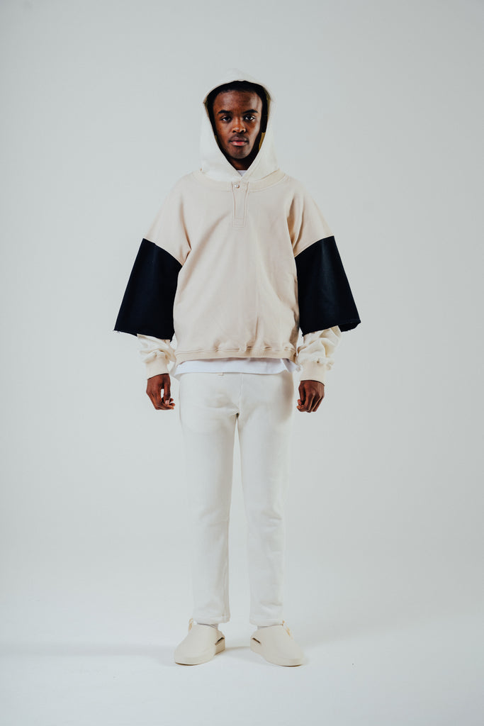 Model wearing a cropped boxy fit Henley sweat in cream and black, styled over a hoodie with white pants.