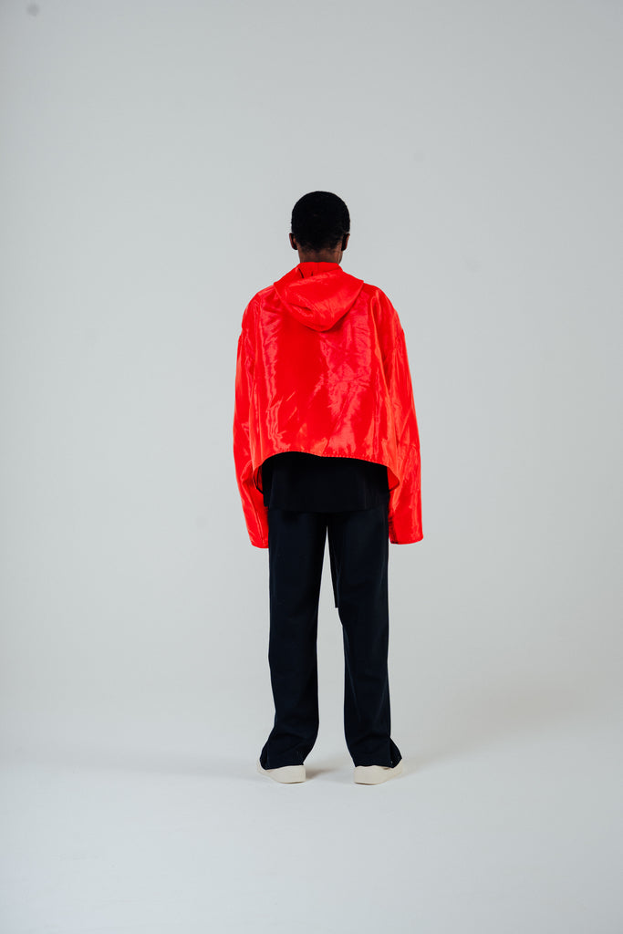 Model in flame red organza hooded zip jacket with cropped body and boxy fit, styled with black pants.