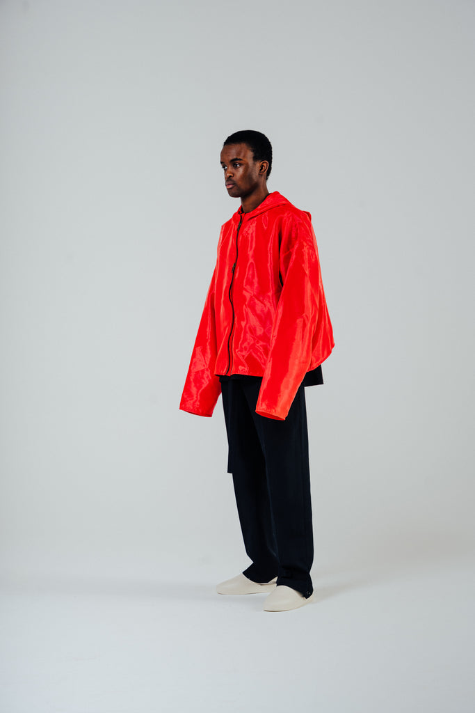 Model wearing a flame red organza hooded zip jacket with a boxy oversized fit and cropped body, styled with dark pants.