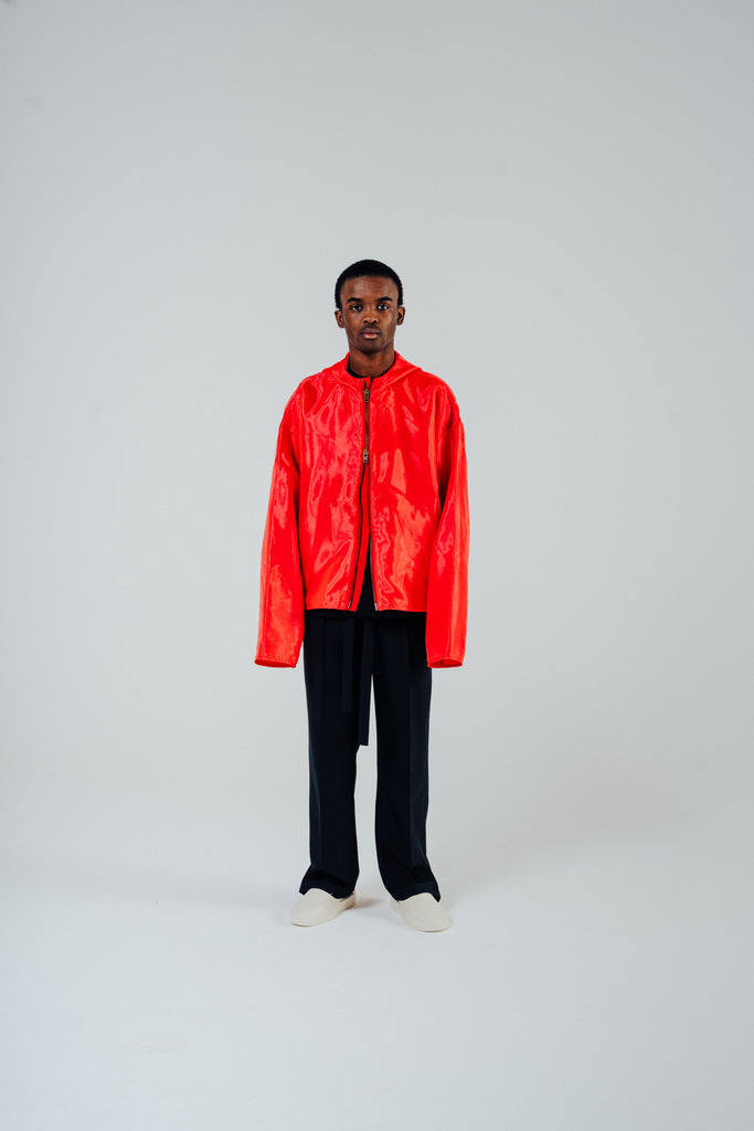 Model wearing a flame red organza hooded zip jacket with a boxy fit, styled with black pants and white shoes.