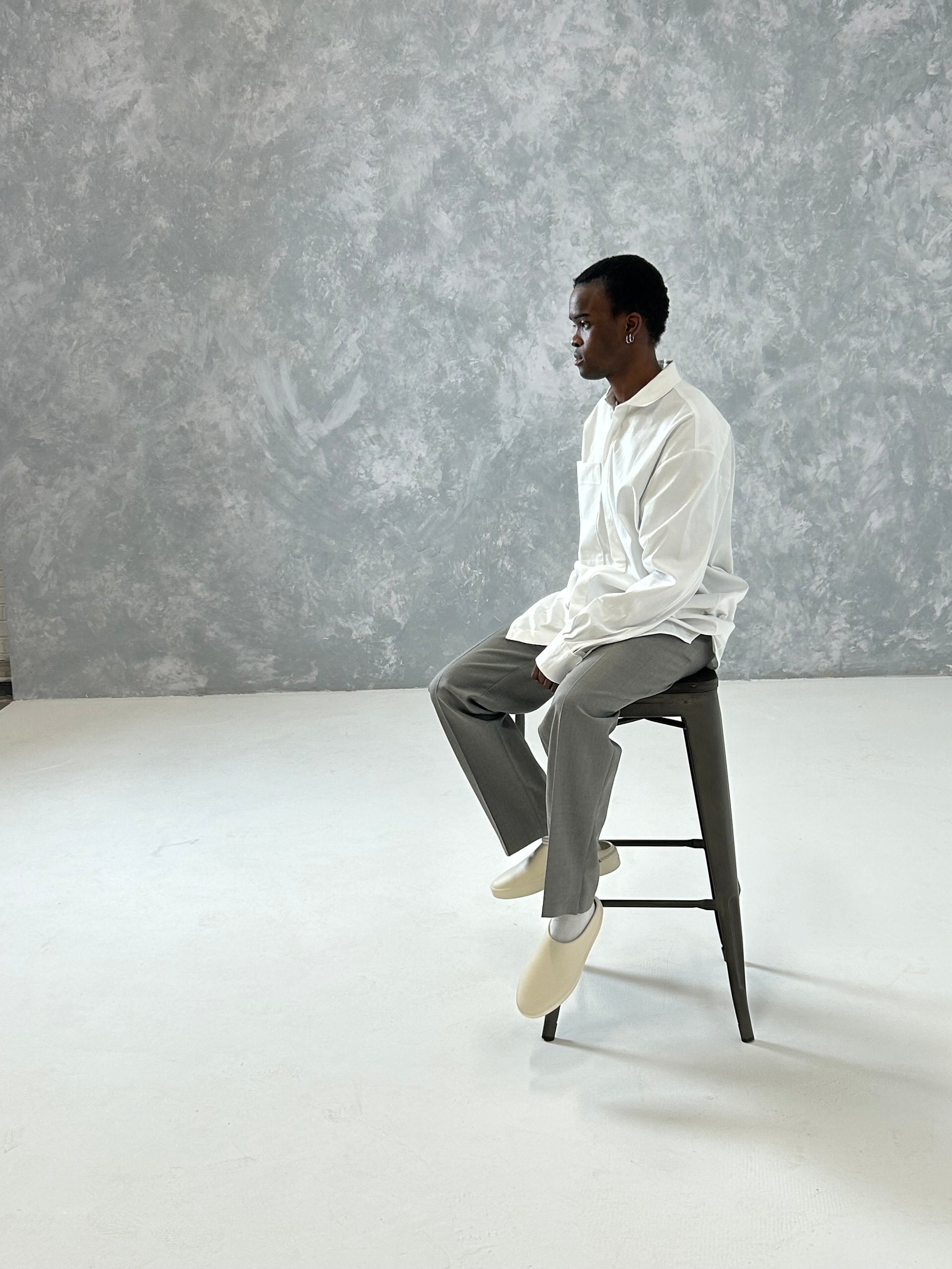 Model wearing an oversized cotton blend shirt in white, seated on a stool against a textured background, showcasing relaxed style.