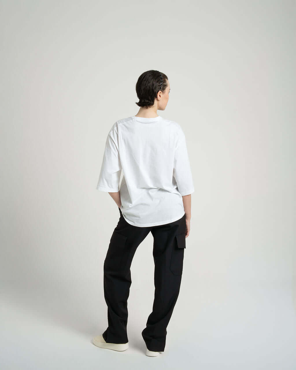 Back view of a model wearing a white boxy cropped tee with slightly lengthened arms and black pants.
