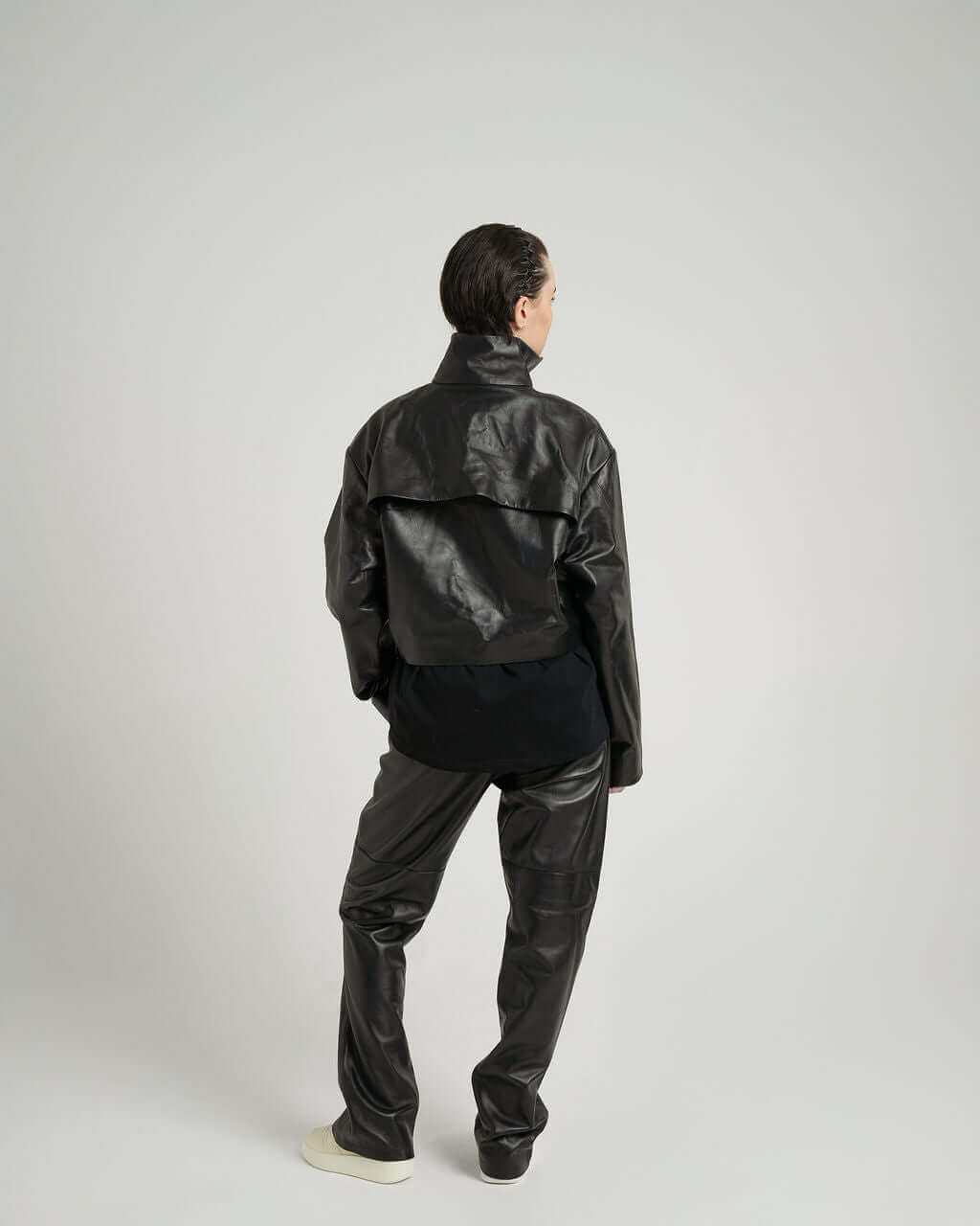 Model wearing a cropped zipped black leather jacket, showcasing the back view with twisted seams and vent details.