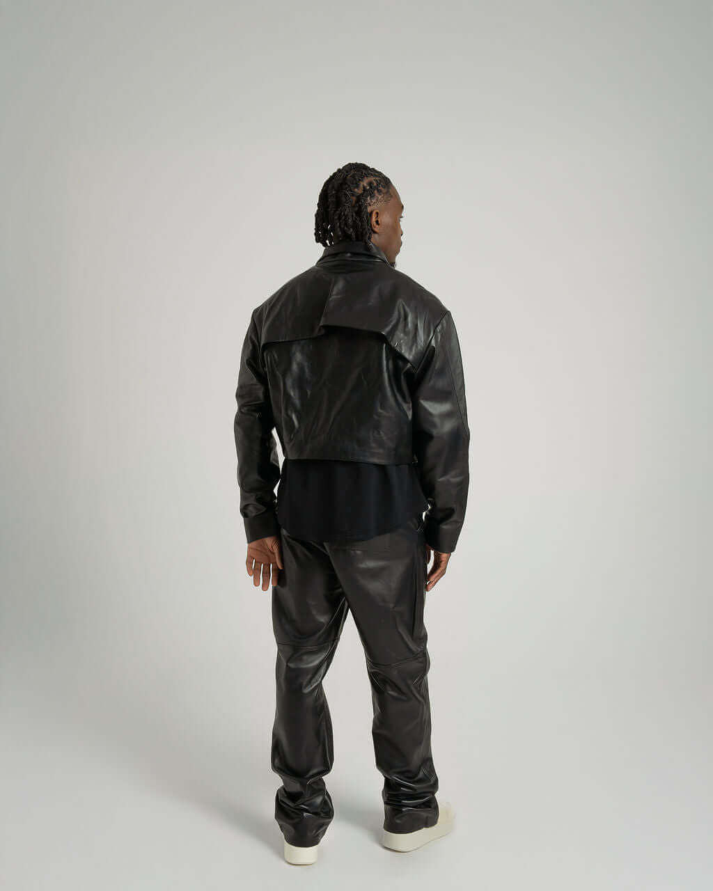 Model wearing a cropped zipped black leather jacket, showing back view and stylish fitted design.