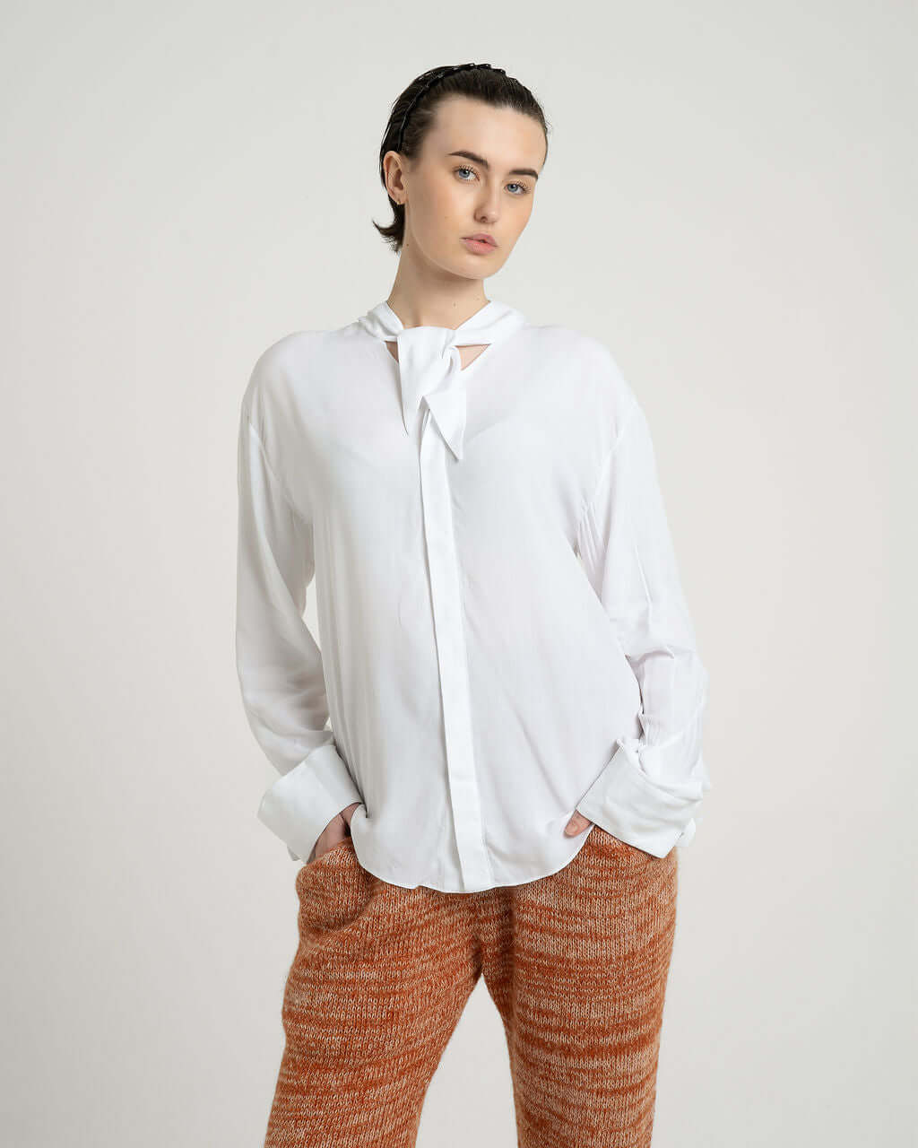 Model wearing a white viscose shirt with neck tie collar and relaxed fit, paired with textured trousers.