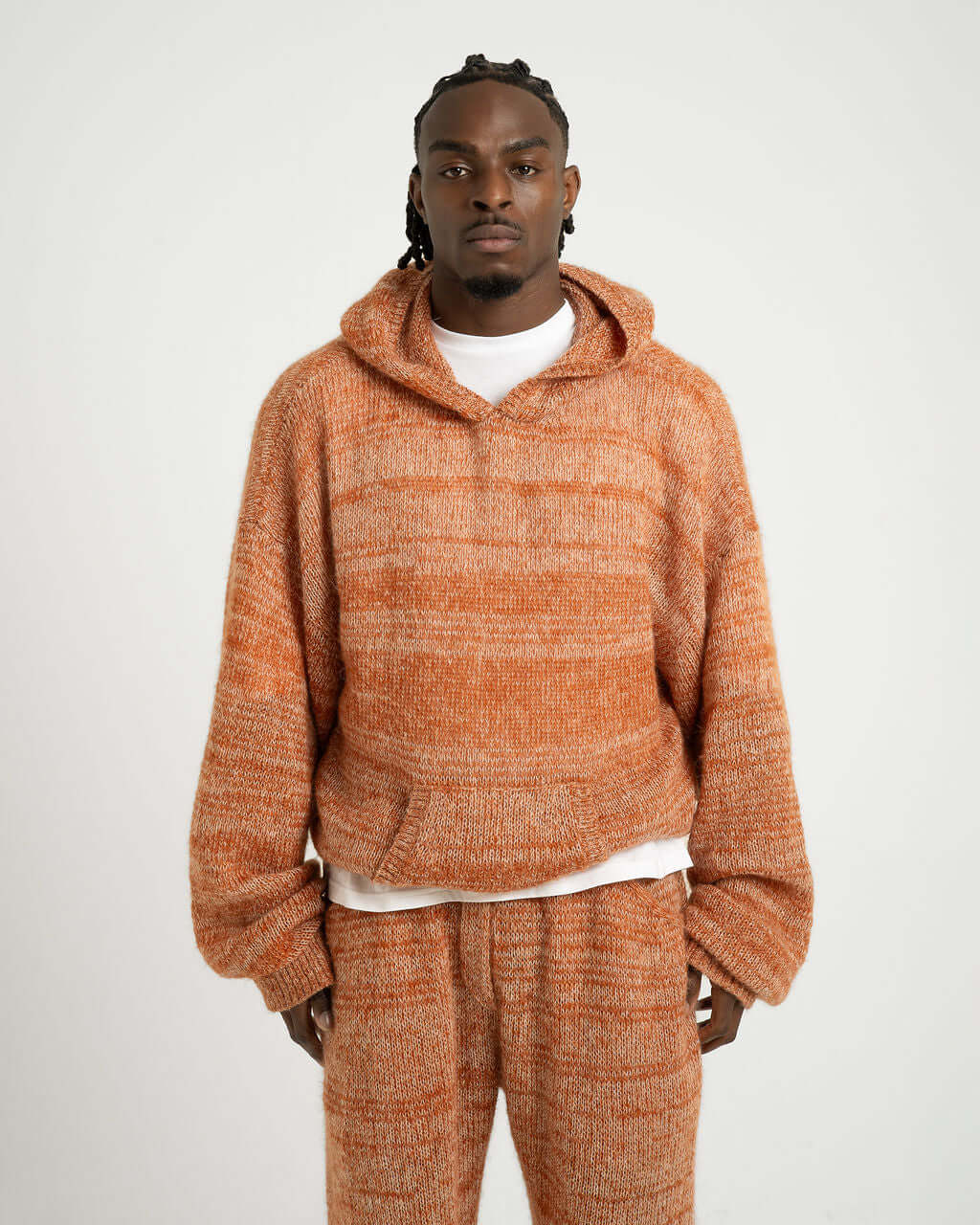 Oversized knitted hoody in orange evening sun, made from mohair blend. Model wearing size L/XL stands against a neutral backdrop.