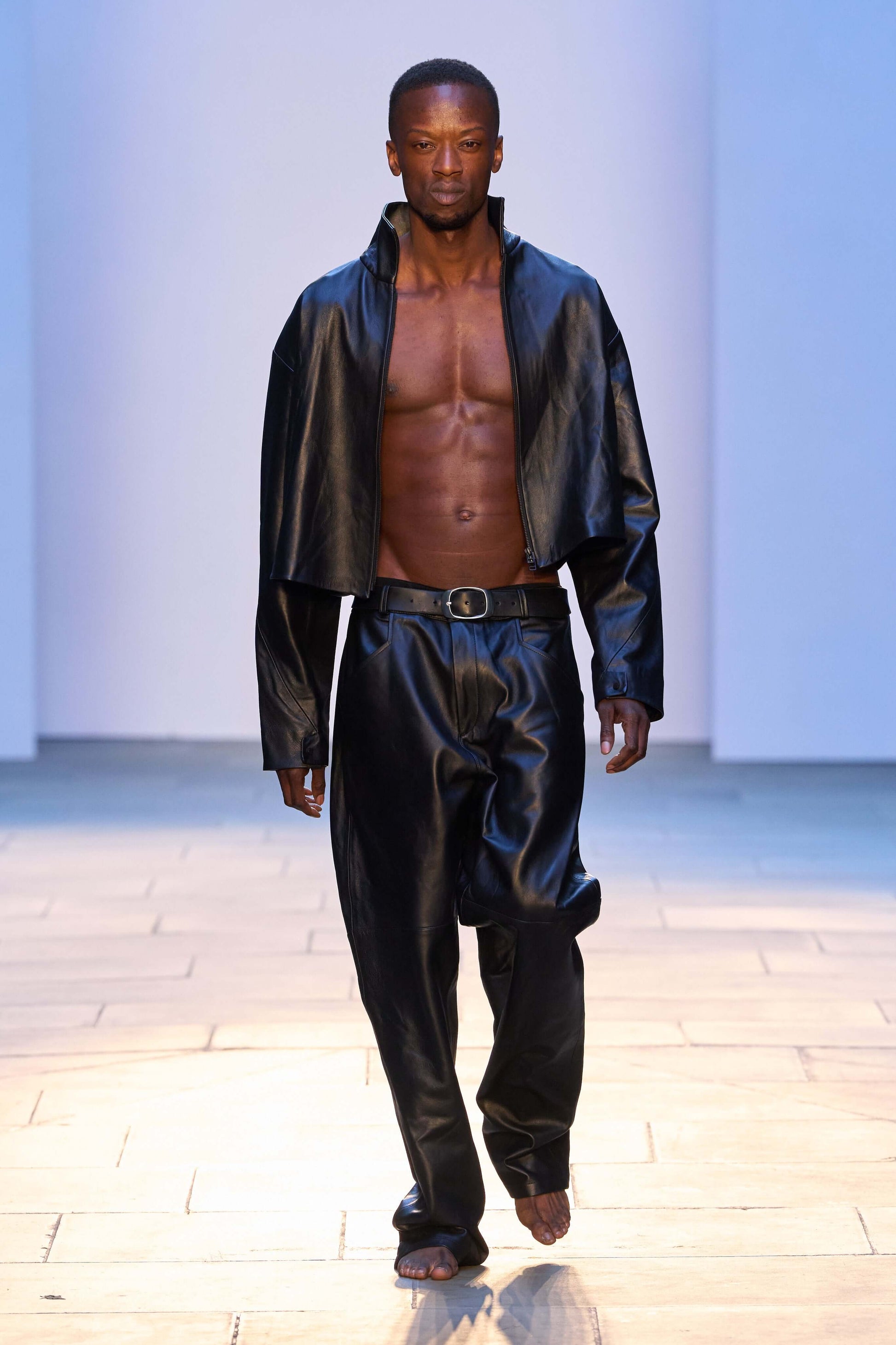 Male model showcasing black leather trousers with a cropped jacket on a runway.